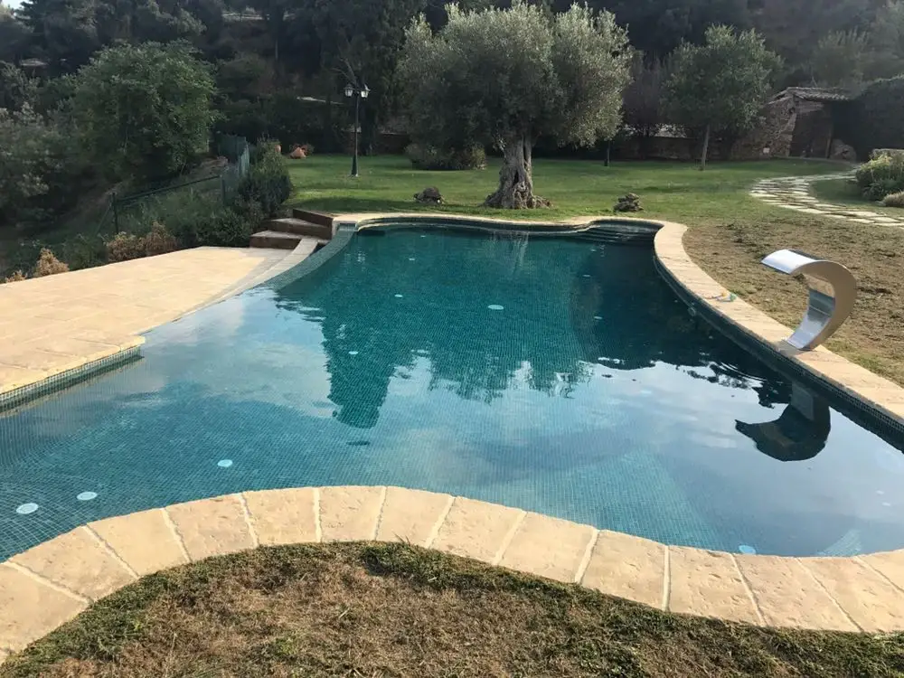 Piscina de formas libres con coronación de piedra clara y una cascada de acero en un jardín con olivos